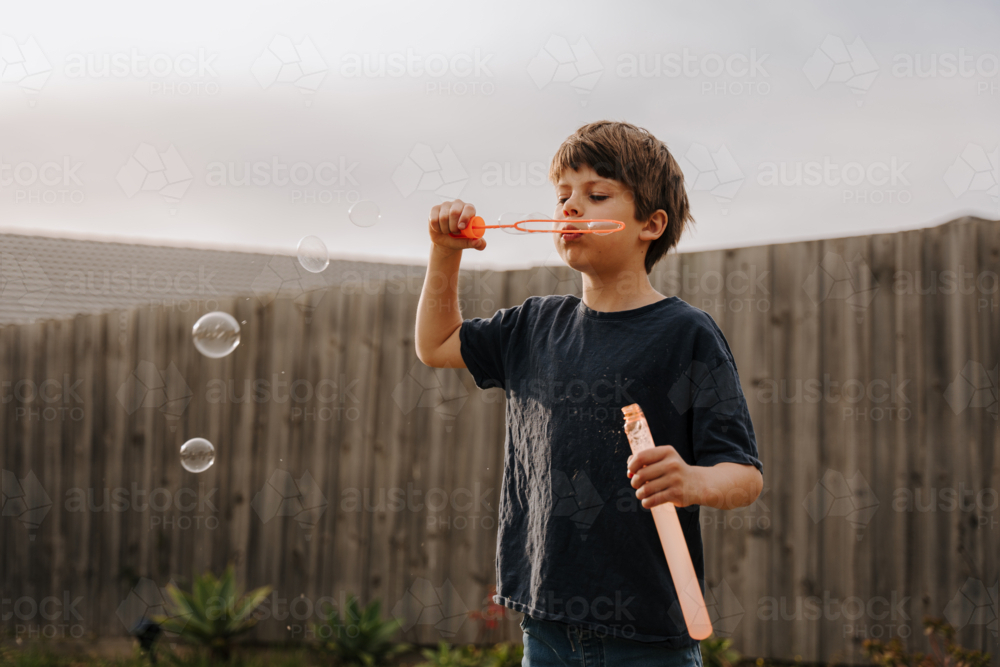 Young boy in black shirt blowing bubbles in their yard. - Australian Stock Image