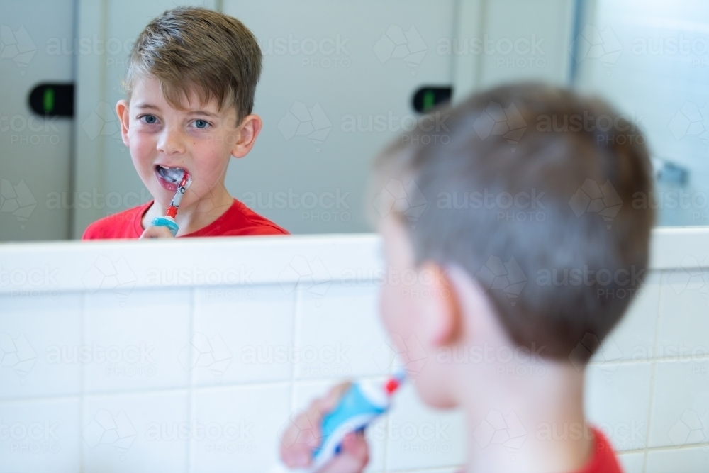 Young boy looking in mirror while cleaning teeth with electric toothbrush - Australian Stock Image