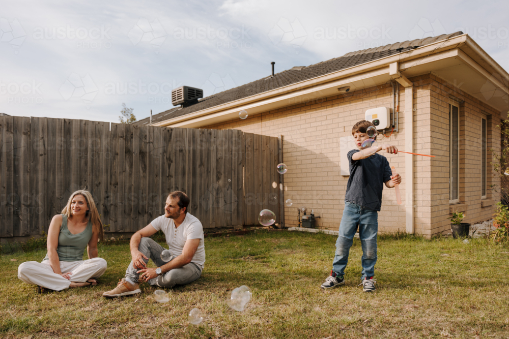 Young boy making and playing with bubbles in their yard while parents sitting on the ground. - Australian Stock Image