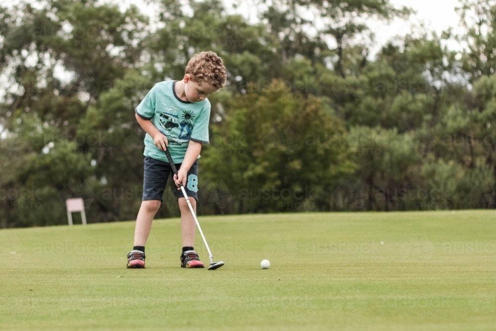 Young boy on green holding golf club putter hitting ball - Australian Stock Image