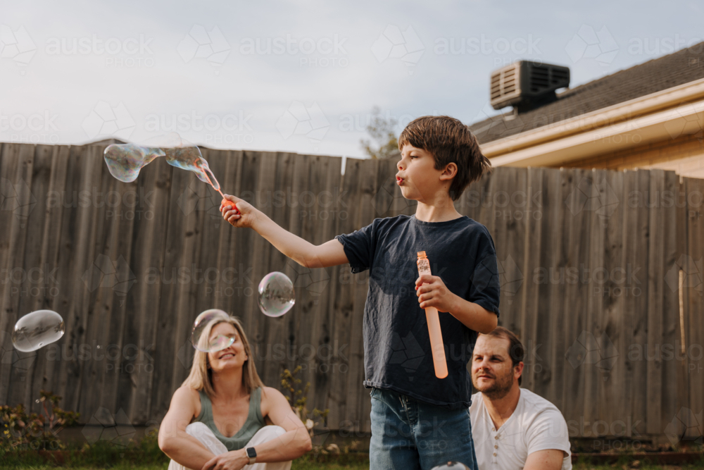 Young boy playing with bubbles in their yard while parents sit on the ground - Australian Stock Image