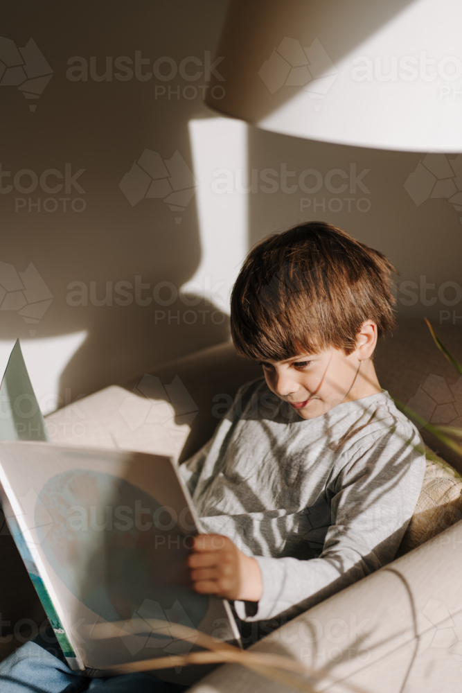 Young boy reading a book on the couch in a sunlit room. - Australian Stock Image