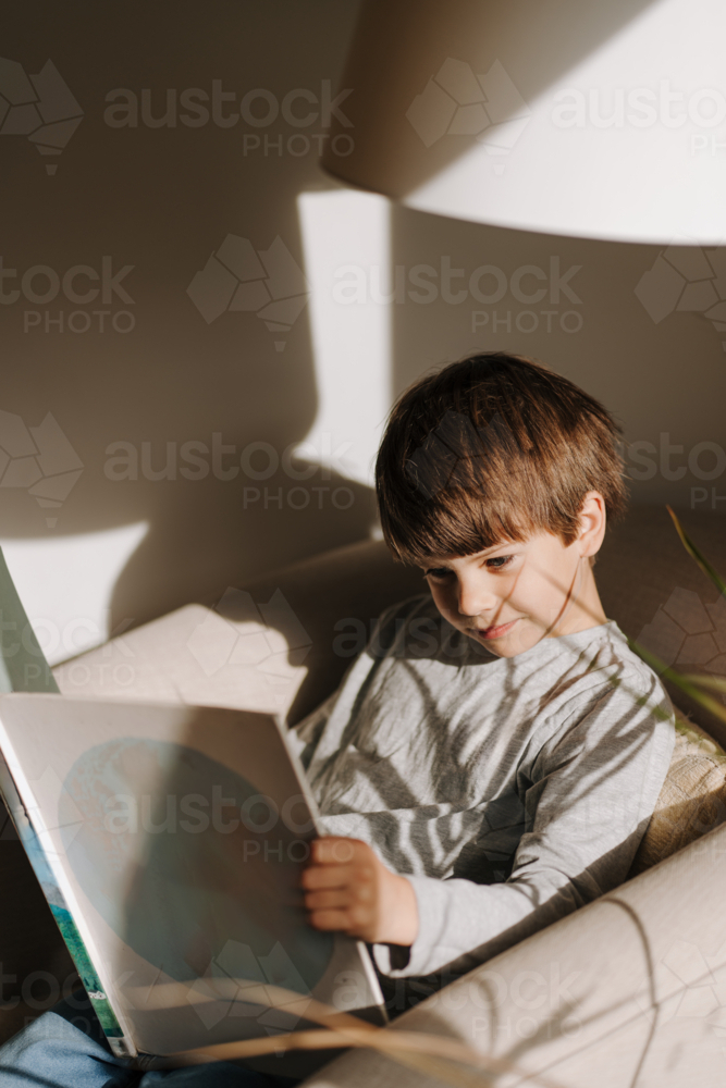 Young boy reading a book on the couch in a sunlit room. - Australian Stock Image