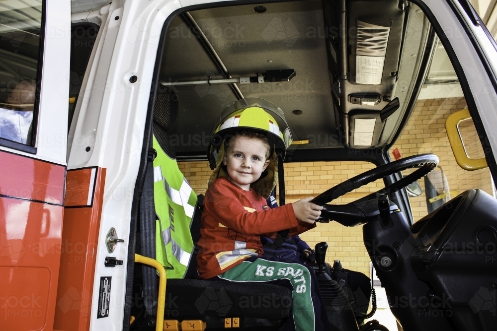 Young boy sitting in fire engine truck wearing helmet with hands on  the steering wheel - Australian Stock Image