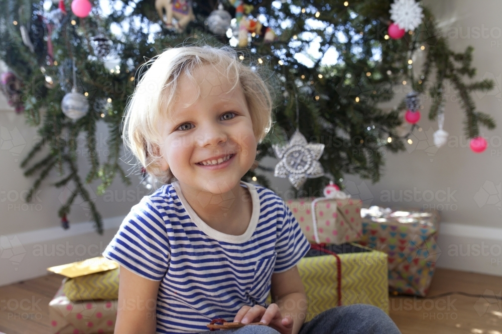 Young boy sitting in front of Christmas tree smiling - Australian Stock Image