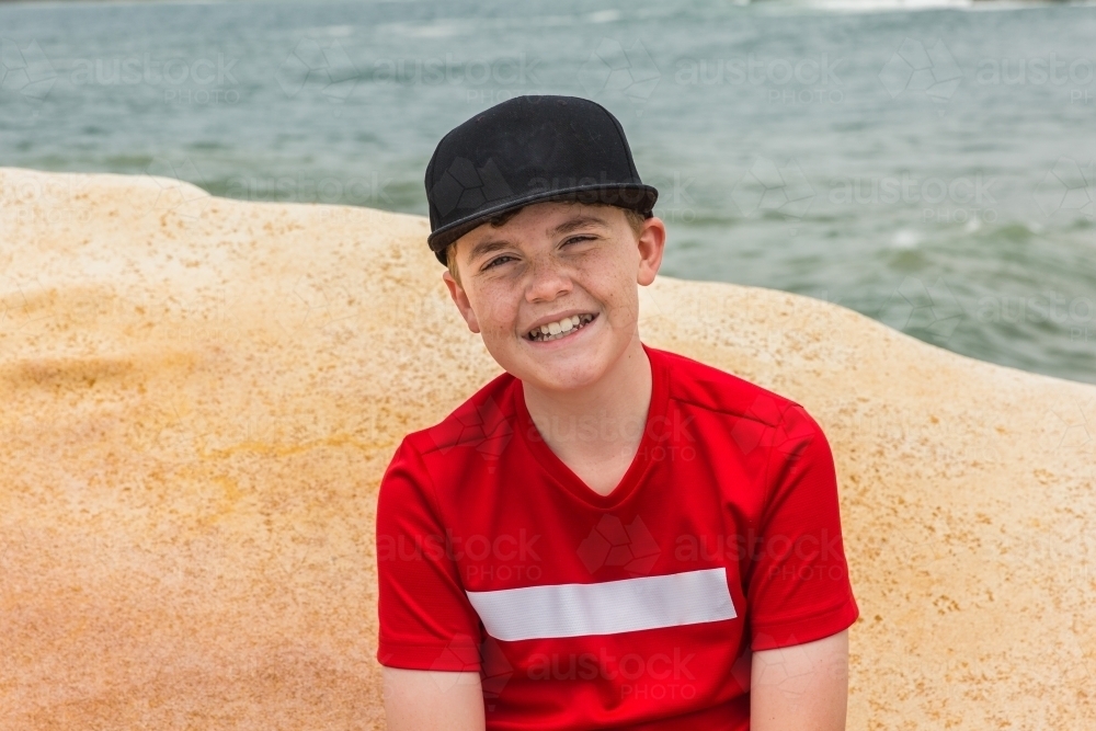 Young boy sitting on rock smiling with ocean behind - Australian Stock Image