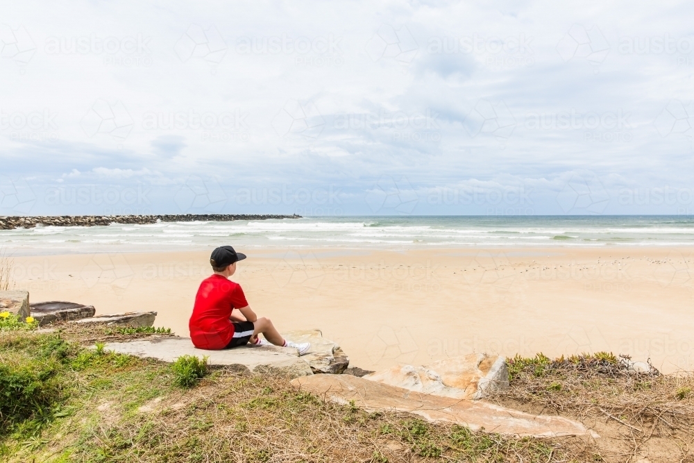 Young boy sitting on rocks looking out over sand and ocean water with rock wall and cloudy skies - Australian Stock Image