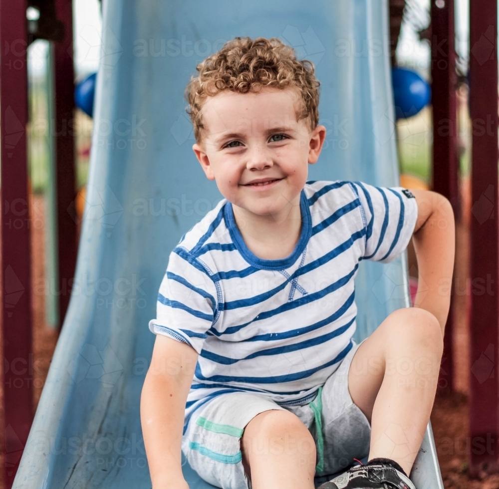 Young boy smiling sitting on end of slippery dip slide at park - Australian Stock Image