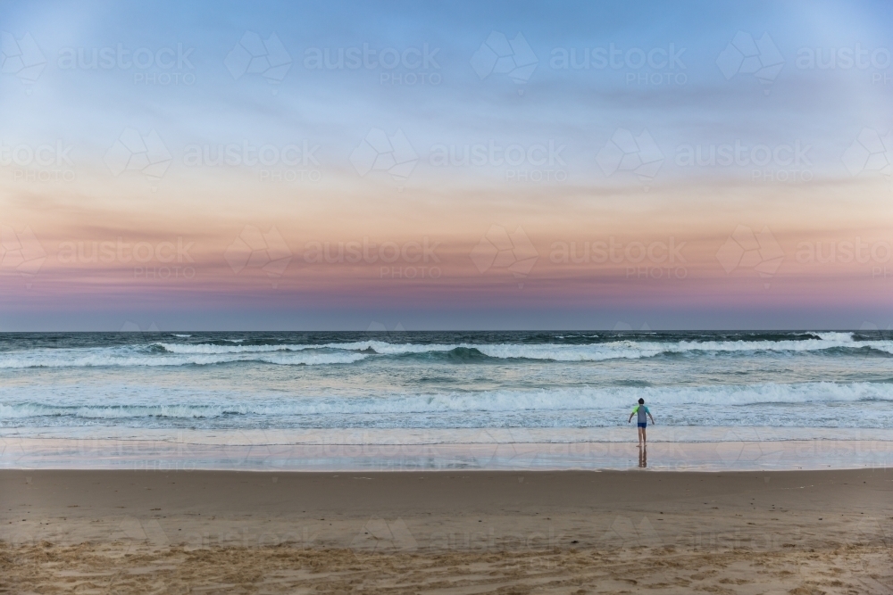 Young boy standing alone on beach looking at waves at sunset - Australian Stock Image