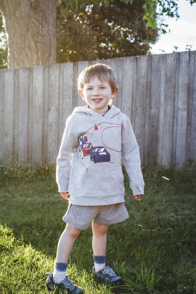 Young boy standing in afternoon sun beside timber fence - Australian Stock Image