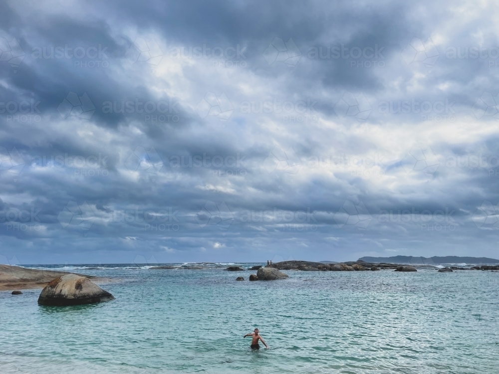 Young boy standing in ocean on overcast day with submerged granite rocks - Australian Stock Image