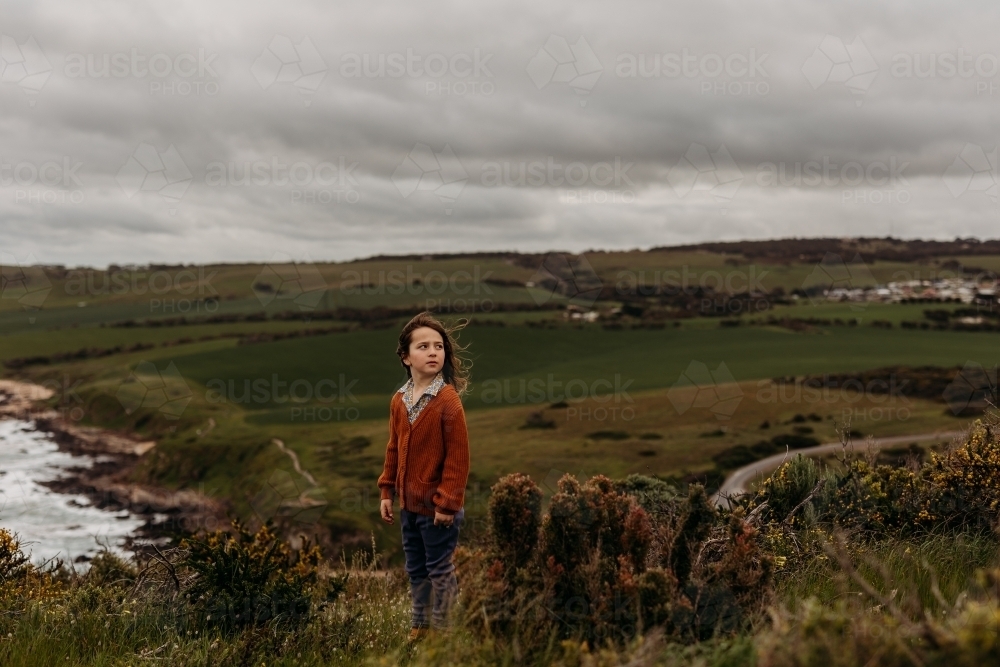 Young boy standing on top of hill with green fields and ocean behind him - Australian Stock Image