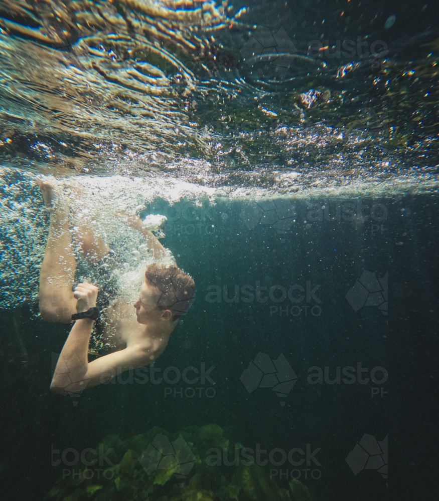 Young boy underwater surrounded by bubbles in clear dark green waters, rocks in background - Australian Stock Image