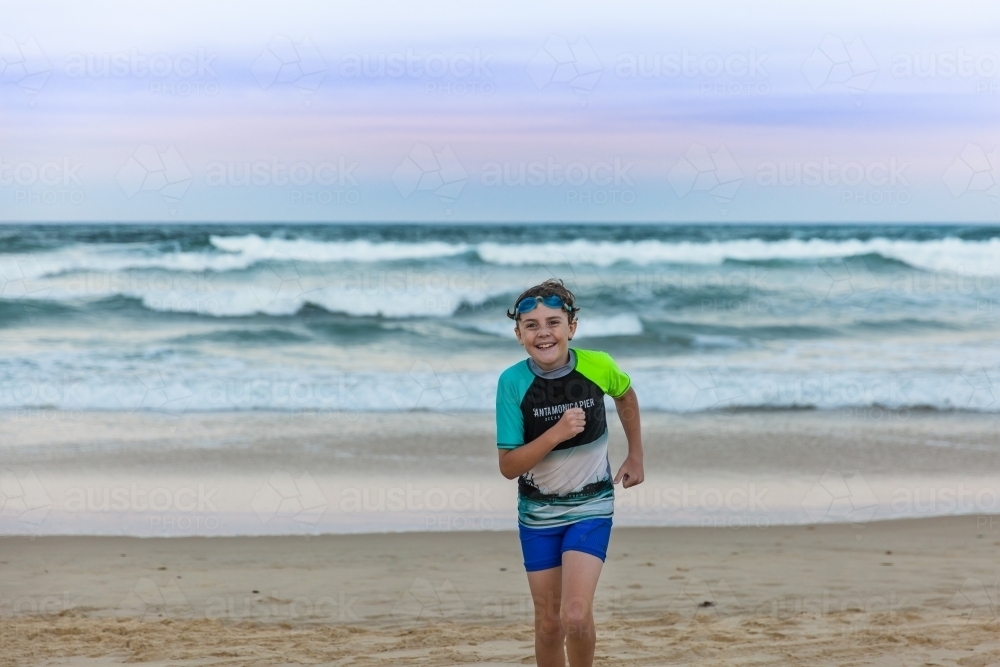 Young boy wearing goggles smiling and jogging on beach with waves at sunset - Australian Stock Image