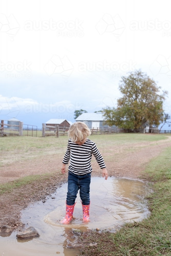 Young boy wearing gumboots standing in muddy puddle on farm - Australian Stock Image