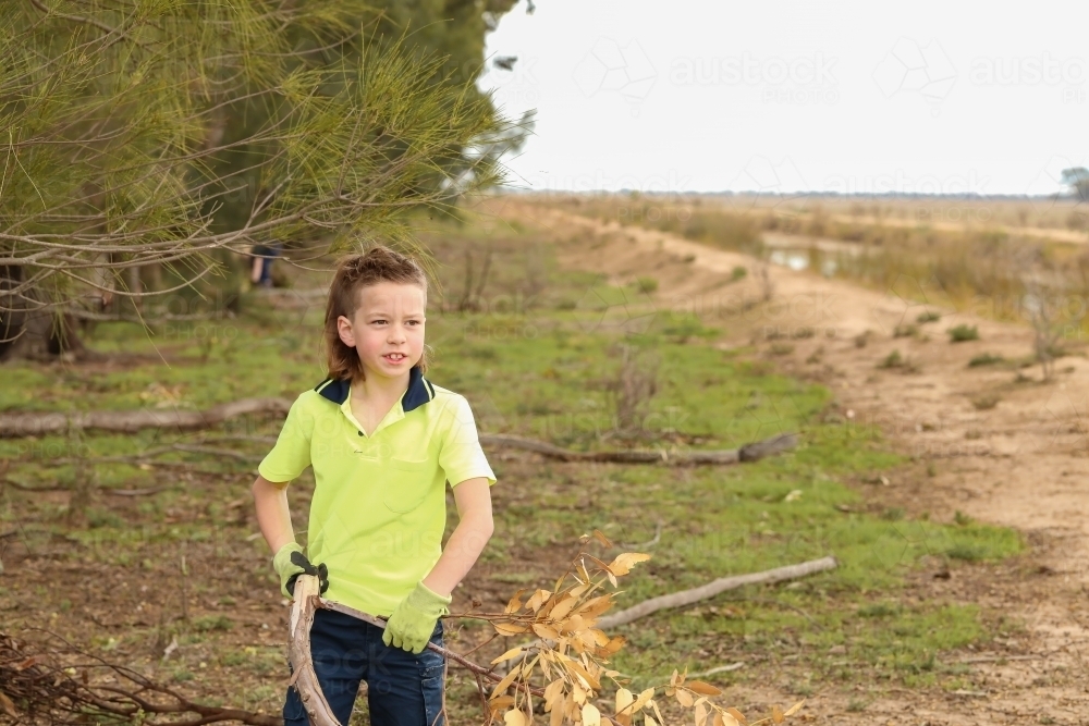 Young boy wearing hi-vis shirt and gloves carrying branch on farm - Australian Stock Image