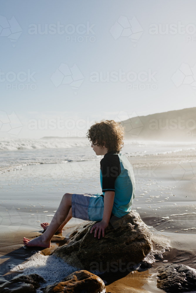 Young boy with curly hair sitting on a rock relaxing at the beach in the afternoon - Australian Stock Image