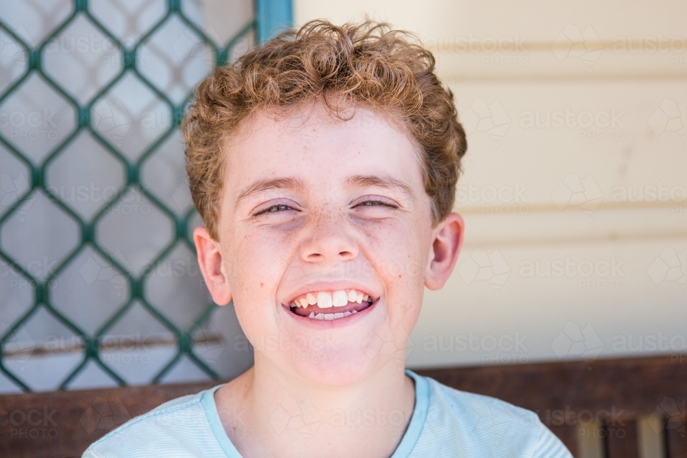 Young boy with curly hair sitting outside house smiling - Australian Stock Image