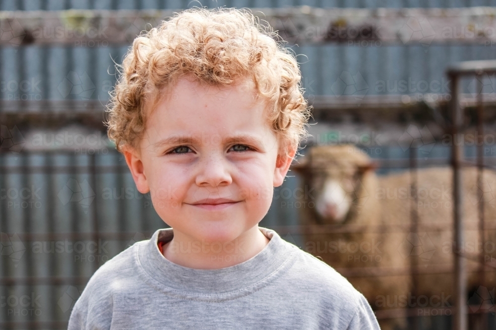 Young boy with curly hair smiling with sheep in background - Australian Stock Image