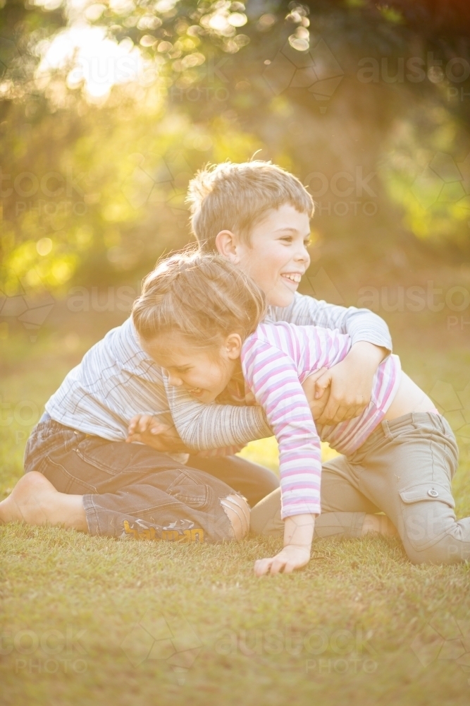 Young brother and sister wrestling on the lawn outside in afternoon light - Australian Stock Image