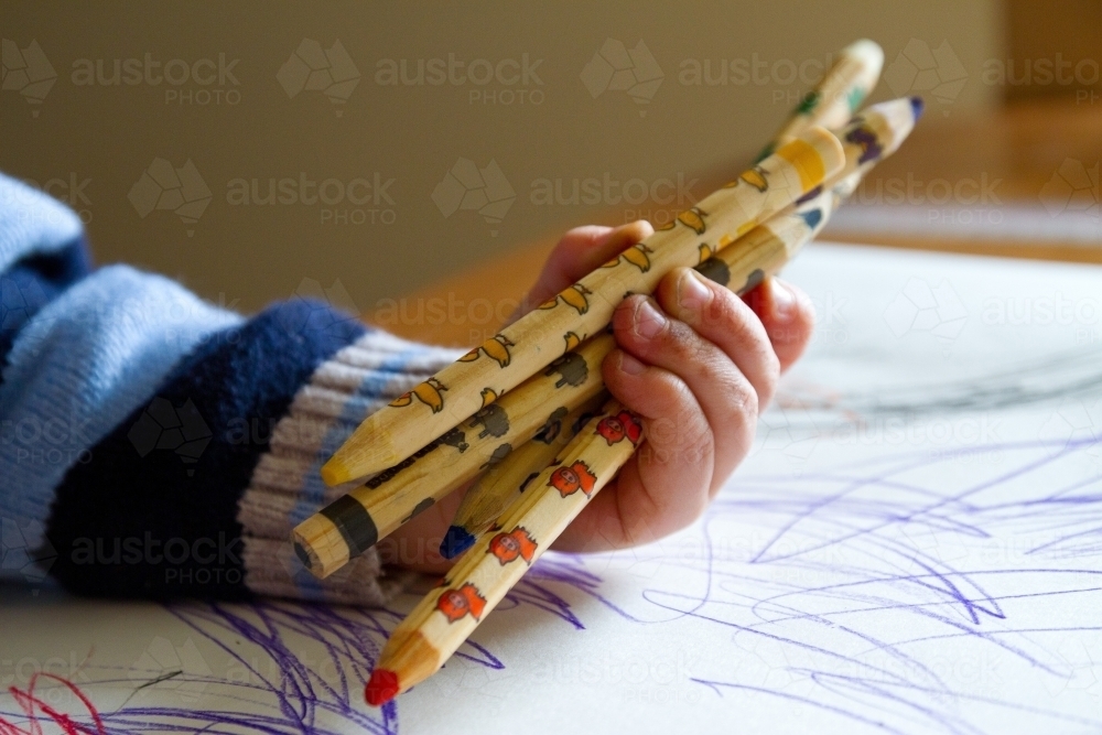 Young child holding colour pencils hovering over drawing artwork on desk - Australian Stock Image