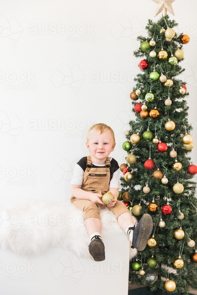 Young child sitting on rug holding bauble next to Christmas tree - Australian Stock Image