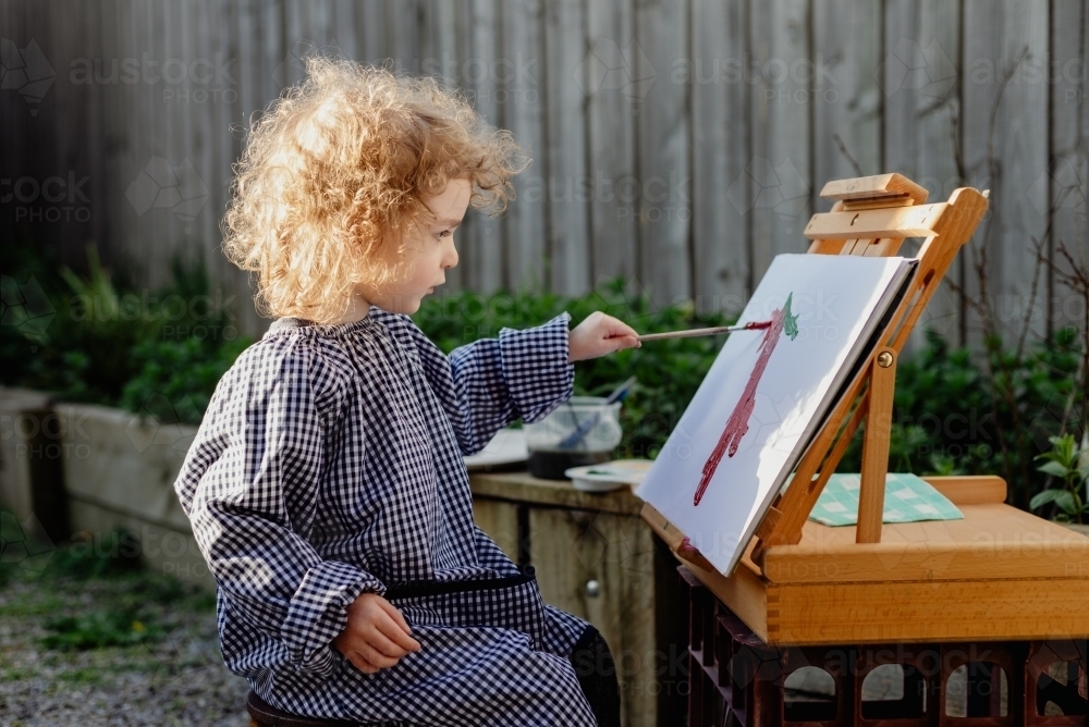 Young child wearing an art smock painting with an easel in the backyard garden at home - Australian Stock Image