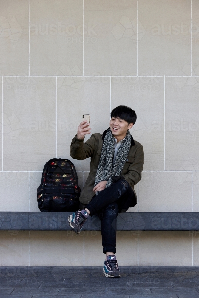 Young Chinese man sitting on bench using mobile phone - Australian Stock Image