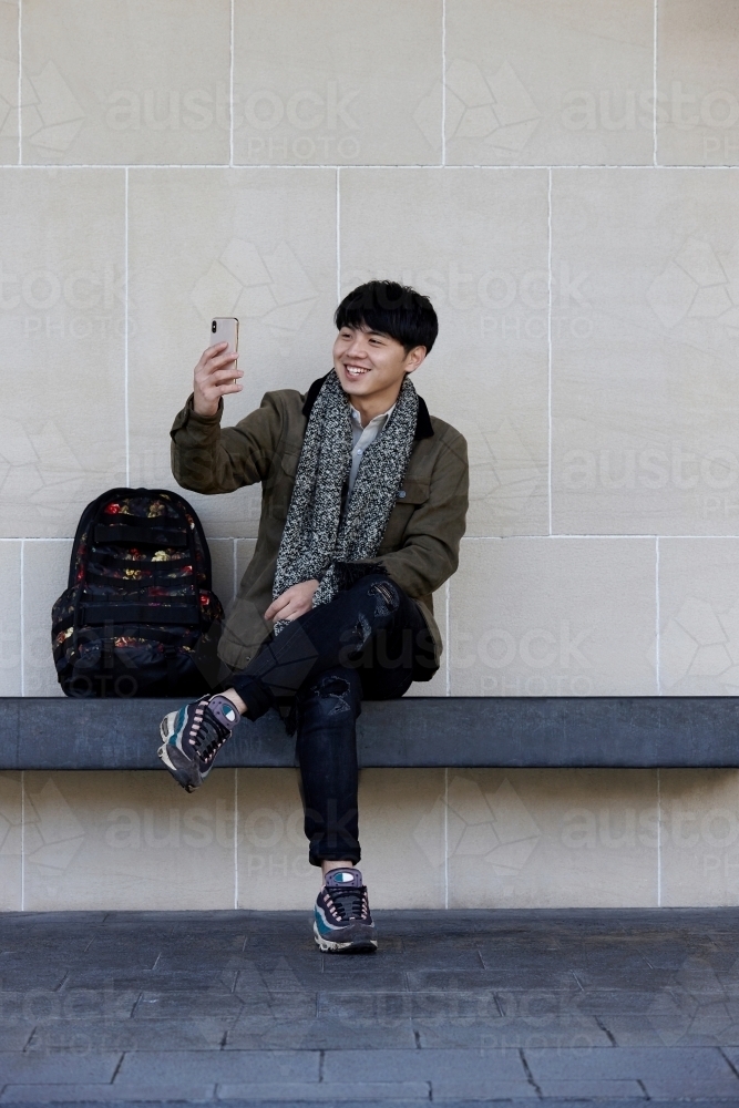 Young Chinese man sitting on bench using mobile phone - Australian Stock Image