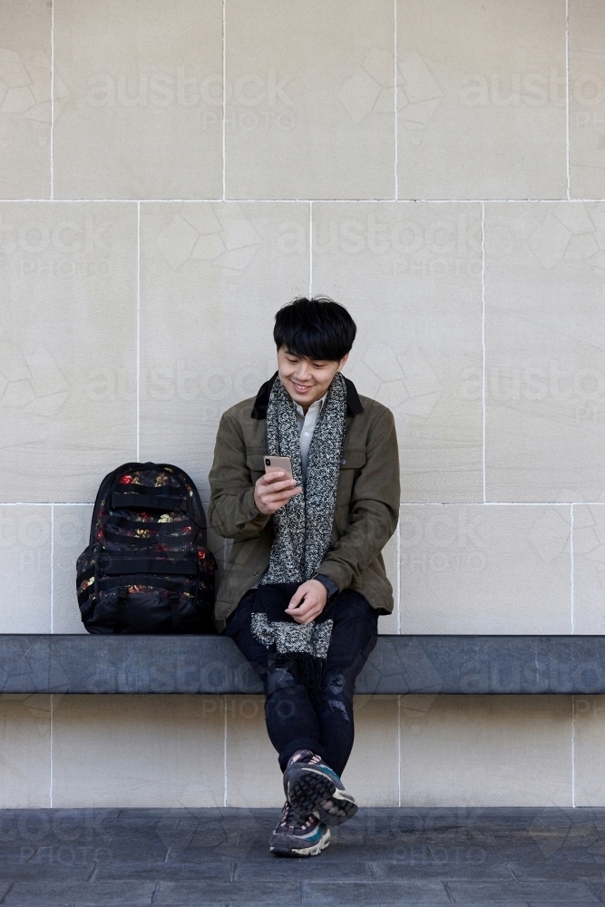 Young Chinese man sitting on bench using mobile phone - Australian Stock Image