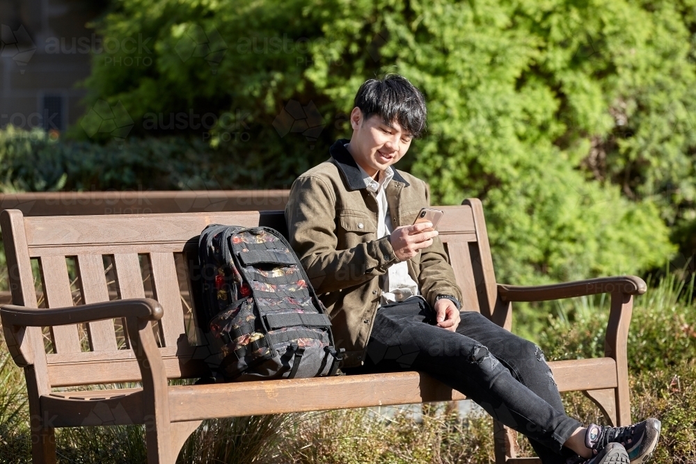 Young Chinese student seated outdoors checking phone with backpack - Australian Stock Image