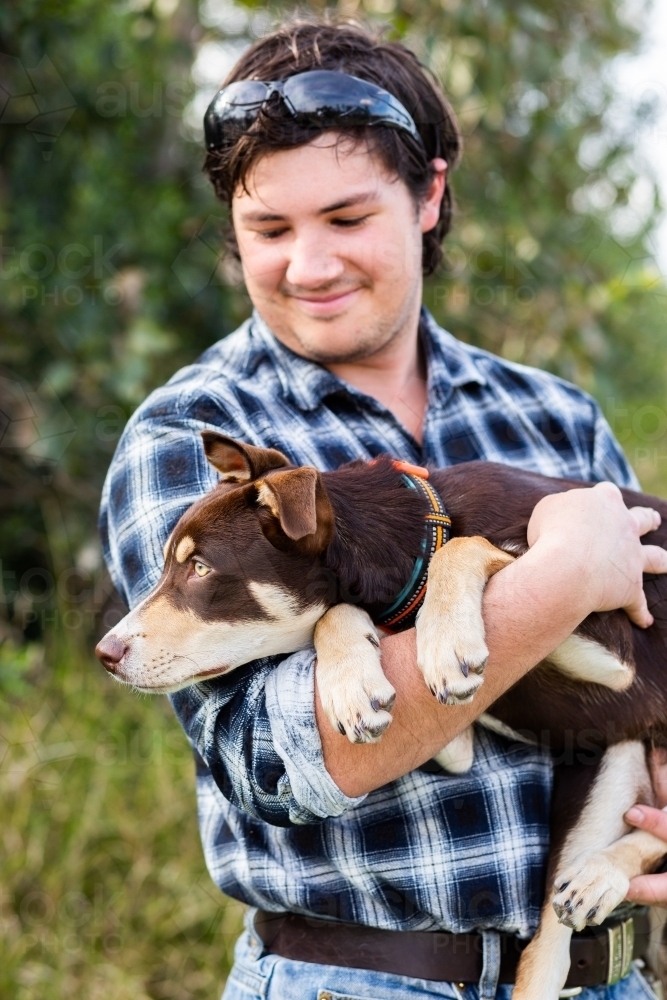 Young country bloke carrying pet Australian Kelpie puppy dog - Australian Stock Image