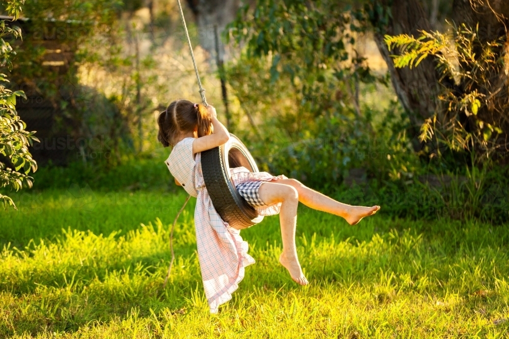 Young country kid in dress swinging on tyre swing in tree outside - Australian Stock Image