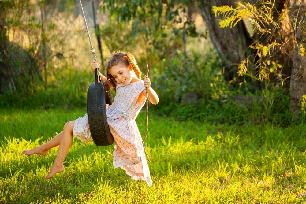 Young country kid in dress swinging on tyre swing in tree outside - Australian Stock Image