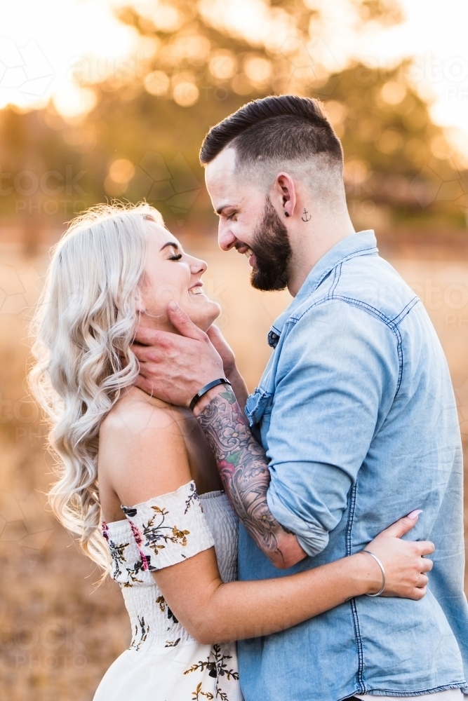 Young couple standing arm in arm while man holds woman's face smiling - Australian Stock Image