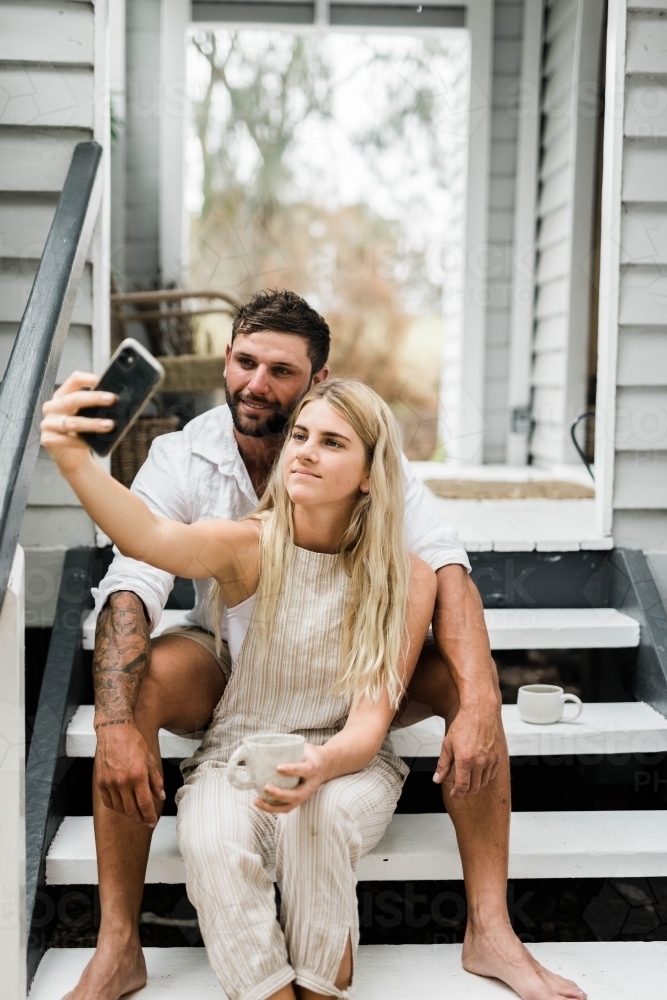 Young couple taking a selfie on the steps of  front porch of weatherboard home - Australian Stock Image