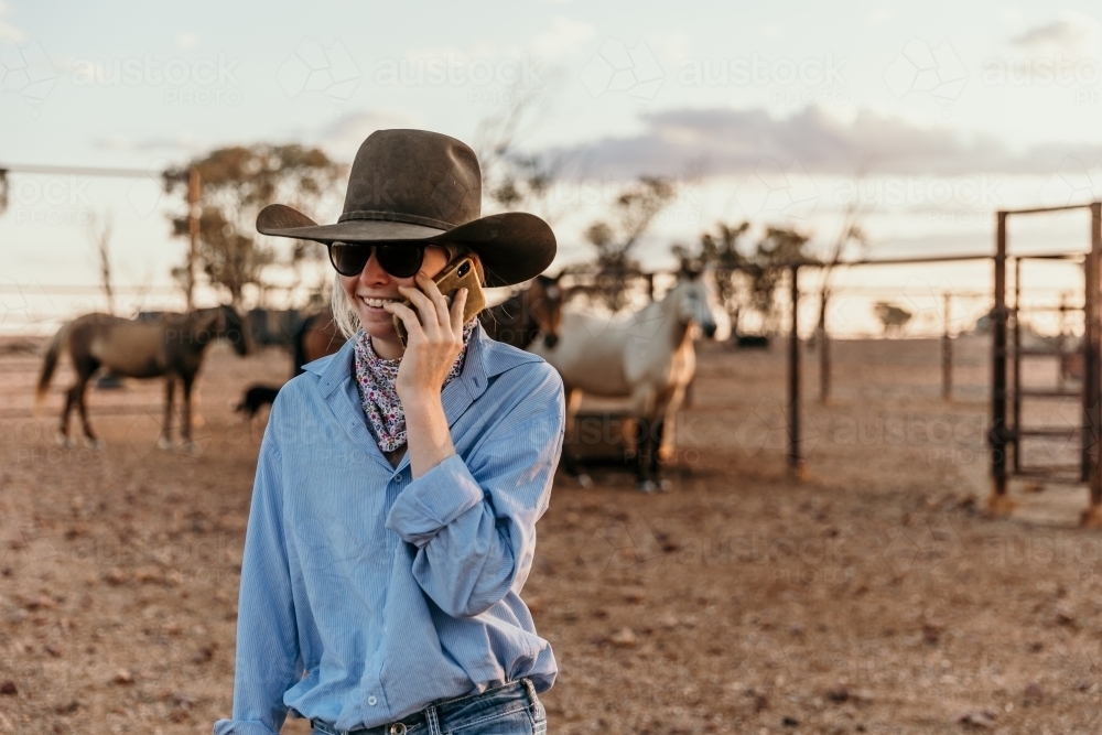 Teenage cowgirl talking on mobile phone in horse yards - Australian Stock Image