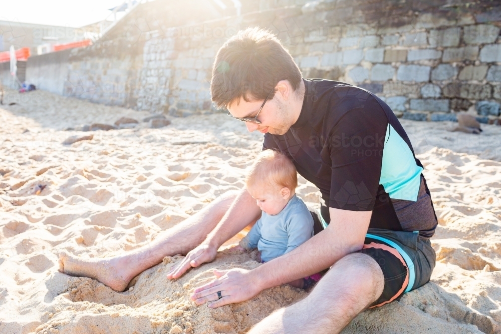 Young dad playing in sand at beach with baby in spring sunlight - Australian Stock Image