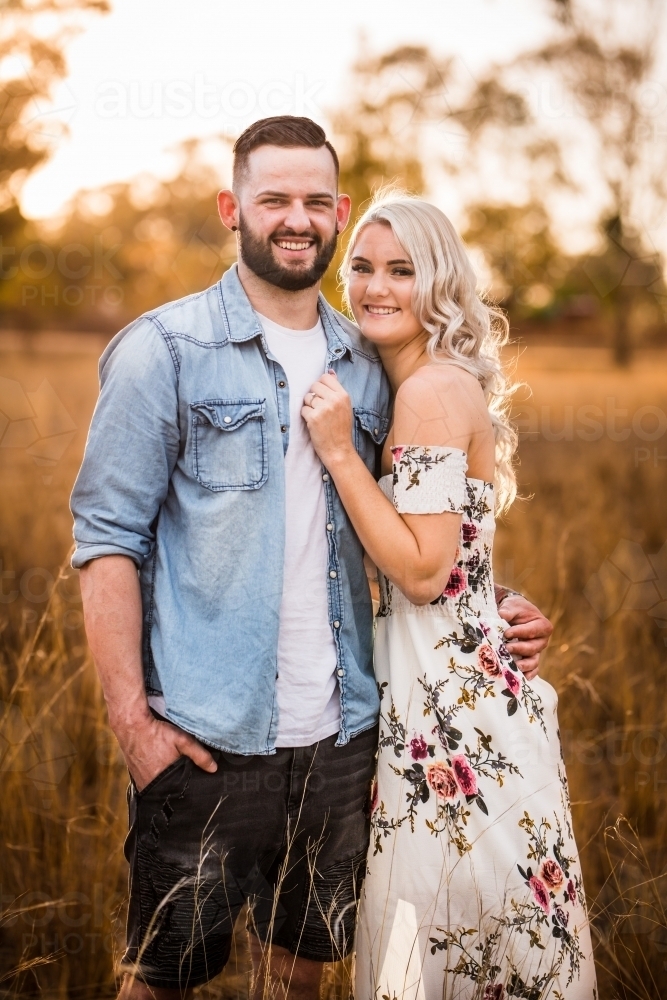 Young engaged couple standing with arms around each other smiling - Australian Stock Image