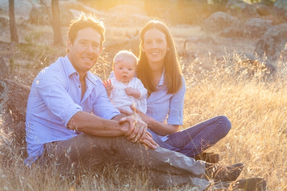 Young family seated in dry grass with sun flare - Australian Stock Image