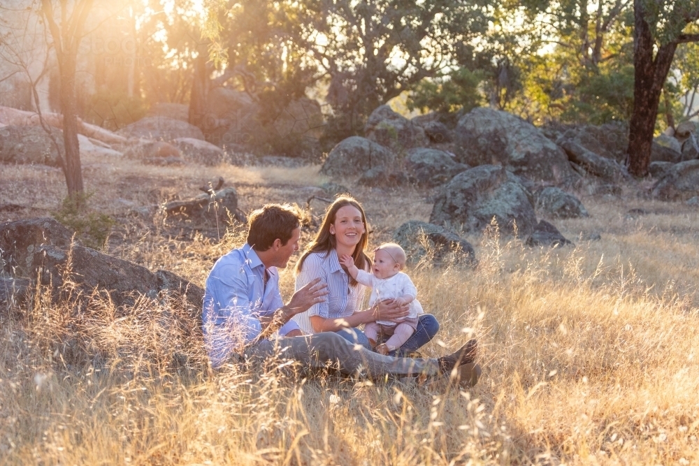 Young family seated in dry grass with sun flare - Australian Stock Image