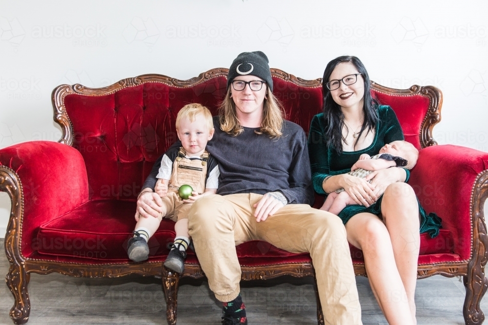 Young family sitting together on red couch smiling holding baby - Australian Stock Image