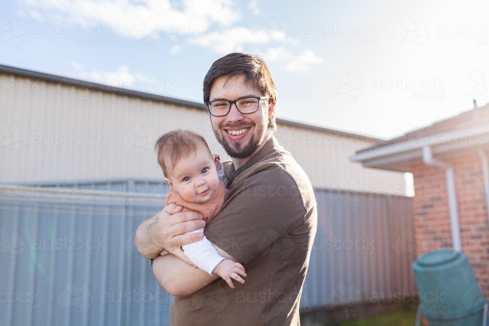 Young father in his twenties holding his baby outside in backyard of home - Australian Stock Image