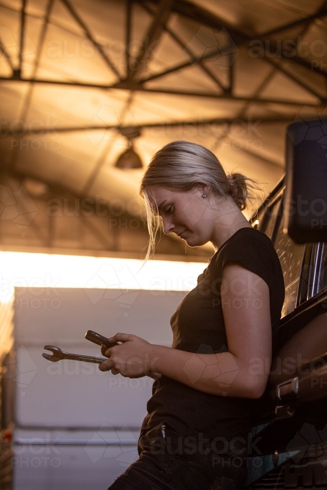 Young female aussie mechanic holding tools and leaning on a car in need of repair in workshop garage - Australian Stock Image