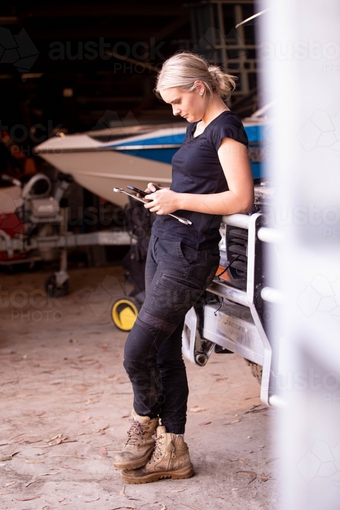 Young female aussie mechanic holding tools and leaning on a car in need of repair in workshop garage - Australian Stock Image