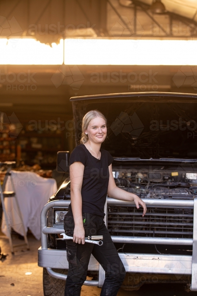 Young female aussie mechanic holding tools and leaning on a car in need of repair in workshop garage - Australian Stock Image