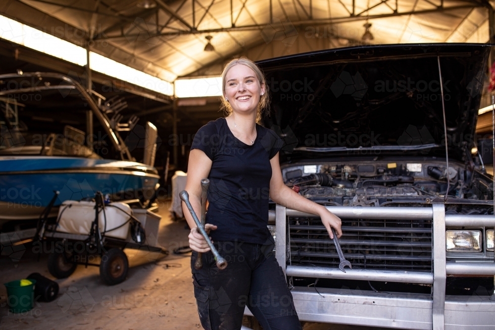 Young female aussie mechanic holding tools and leaning on a car in need of repair in workshop garage - Australian Stock Image