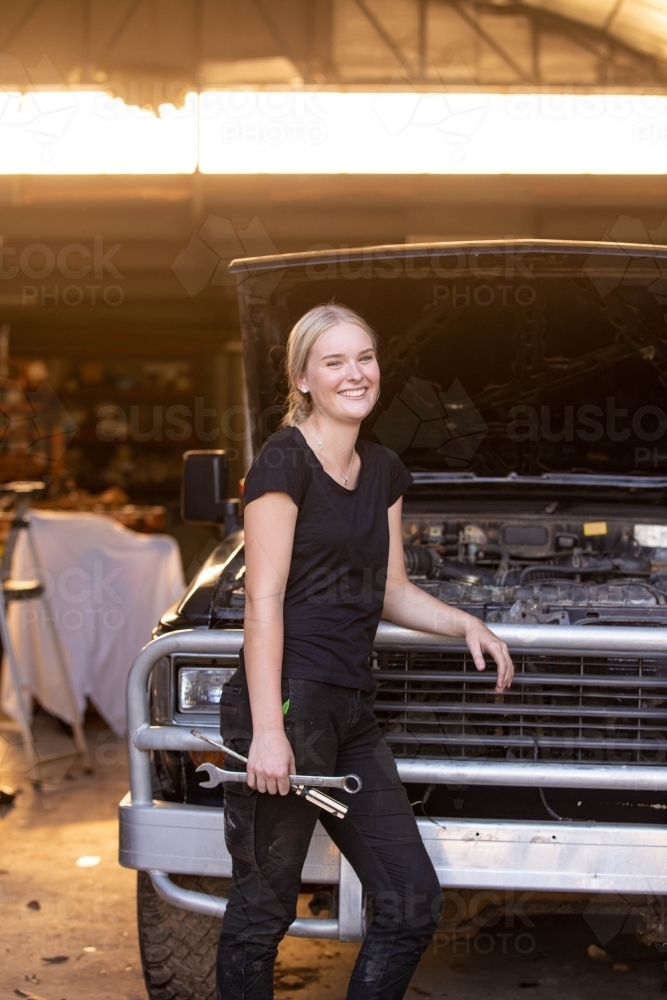 Young female aussie mechanic holding tools and leaning on a car in need of repair in workshop garage - Australian Stock Image