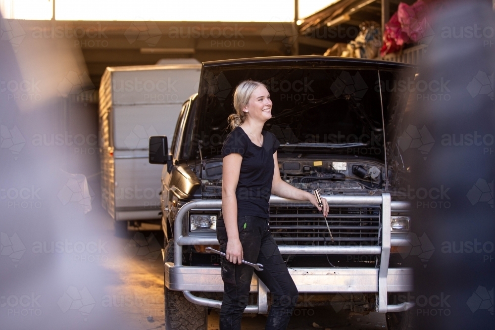 Young female aussie mechanic holding tools and leaning on a car in need of repair in workshop garage - Australian Stock Image