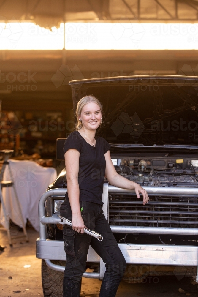 Young female aussie mechanic leaning on a car in need of repair in workshop holding a wrench - Australian Stock Image